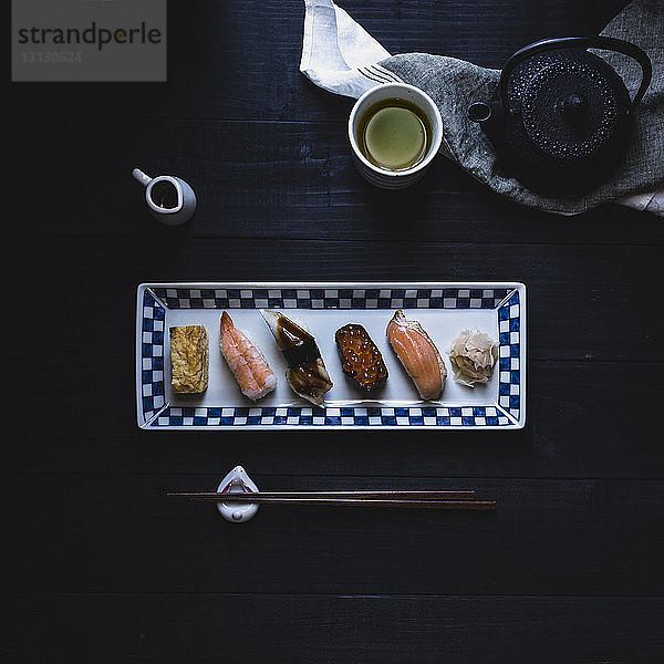 Draufsicht auf verschiedene Sushi in Tellerform mit Essstäbchen und grünem Tee auf schwarzem Tisch