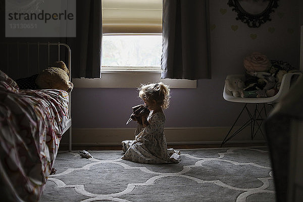 Seitenansicht eines Mädchens  das auf einem Teppich sitzend gegen das Fenster im Schlafzimmer spielt