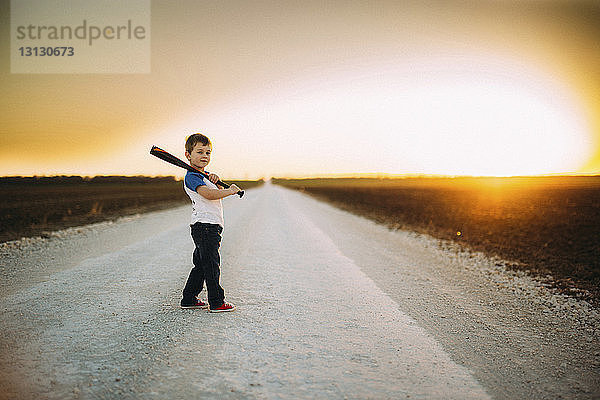 Porträt eines Jungen  der einen Baseballschläger hält  während er bei Sonnenuntergang auf der Straße inmitten des Feldes gegen den Himmel steht