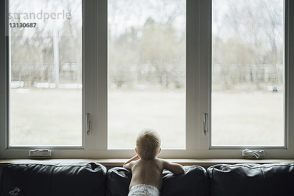 Rückansicht eines Jungen ohne Hemd  der durch das Fenster schaut  während er zu Hause auf dem Sofa steht