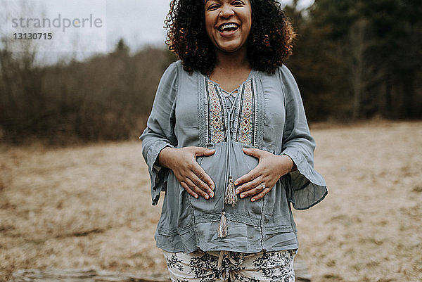 Porträt einer glücklichen schwangeren Frau  die den Bauch berührt  während sie im Wald steht