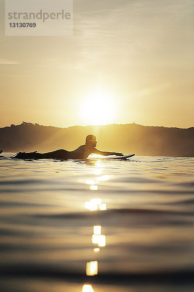 Seitenansicht einer Surferin  die bei Sonnenuntergang auf einem Surfbrett im Meer liegt