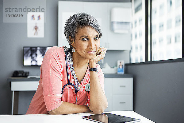 Porträt einer selbstbewussten Ärztin  die in der Klinik am Schreibtisch lehnt