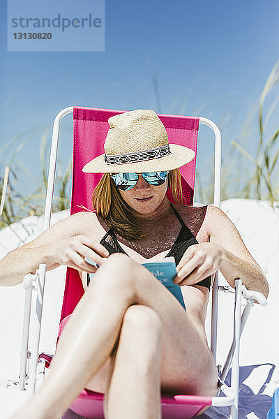 Junge Frau liest bei sonnigem Wetter beim Sonnenbaden am Strand ein Buch