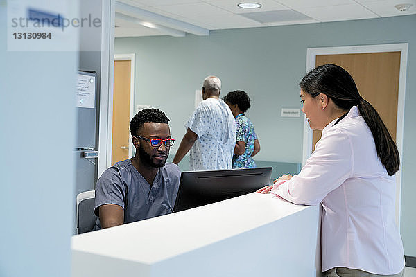 Arzt im Gespräch mit Krankenpfleger am Krankenhausempfang  während Kollege mit Patient im Hintergrund geht