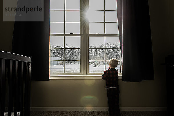 Rückansicht eines Jungen  der durch ein Fenster schaut  während er im Winter zu Hause steht