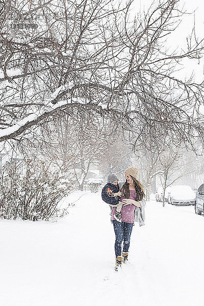 Schwangere Mutter trägt einen Sohn  während sie bei Schneefall auf verschneitem Feld in der Stadt läuft