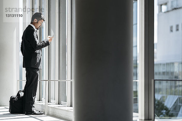 Seitenansicht eines Geschäftsmannes  der ein Smartphone benutzt  während er im Büro am Fenster steht