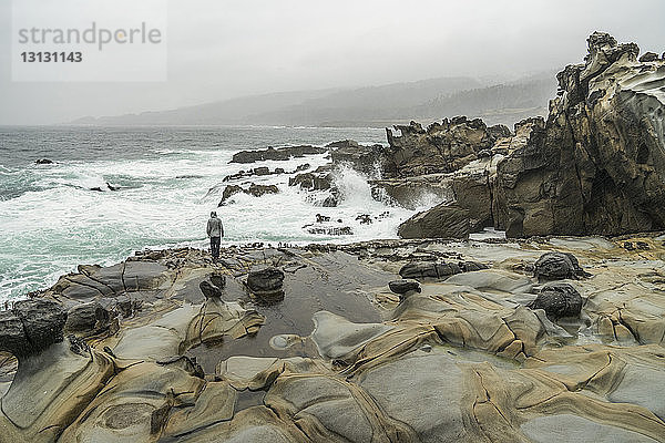Hochwinkelaufnahme eines Mannes  der bei nebligem Wetter auf Felsen am Strand gegen den Himmel steht