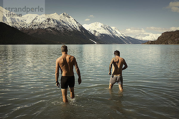 Rückansicht der hemdlosen männlichen Freunde  die in den Kenai-See vor schneebedeckten Bergen gehen