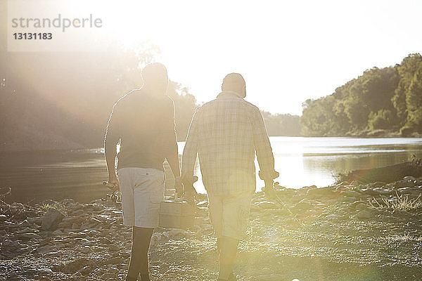 Rückansicht von männlichen Freunden mit Angelruten  die bei Sonnenschein auf Felsen am See spazieren