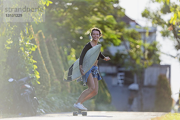 Sorglose Frau fährt Skateboard  während sie bei Sonnenschein das Surfbrett auf der Straße hält