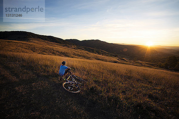 Männlicher Athlet mit Fahrrad  der bei Sonnenuntergang auf einem Grasfeld gegen den Himmel sitzt