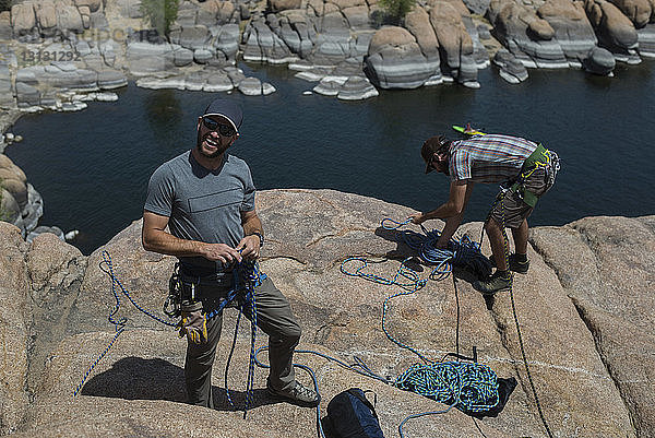 Hochwinkelaufnahme von Freunden  die bei Sonnenschein Kletterseile binden  während sie auf einer Felsformation über dem See stehen