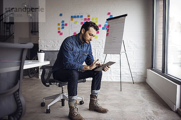 Seriöser Geschäftsmann benutzt Tablet  während er in einem kreativen Büro auf einem Stuhl sitzt