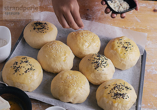 Abgehackte Hände eines Jungen  der zu Hause in der Küche Sesamkörner auf Brötchen schmiert