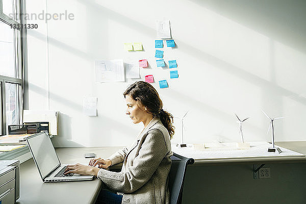 Seitenansicht einer Geschäftsfrau  die einen Laptop benutzt  während sie an auf dem Schreibtisch angeordneten Modellen von Windkraftanlagen arbeitet