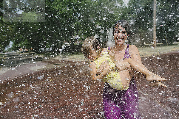 Verspielte Mutter  die ihre Tochter austrägt  während sie sich im Springbrunnen im Park vergnügt