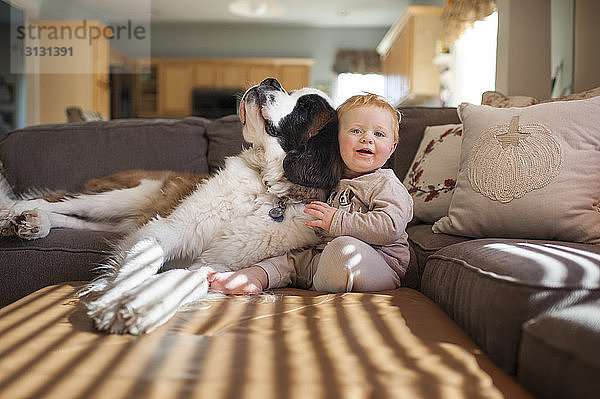 Porträt eines süßen kleinen Jungen  der mit seinem Hund auf der Couch im heimischen Wohnzimmer spielt