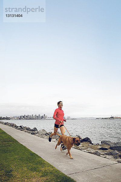 Sportlerin rennt mit Hund auf Fußpfad am Meer gegen klaren Himmel