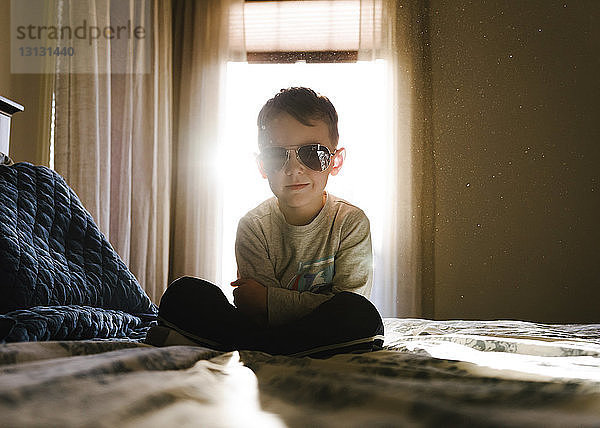 Bildnis eines Jungen mit verschränkten Armen und Sonnenbrille  der zu Hause auf dem Bett sitzt