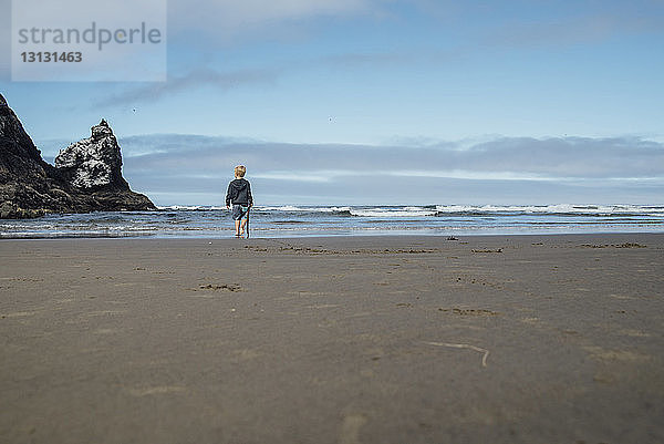 Junge in voller Länge mit Stock in der Hand  während er auf Sand am Strand gegen den Himmel läuft