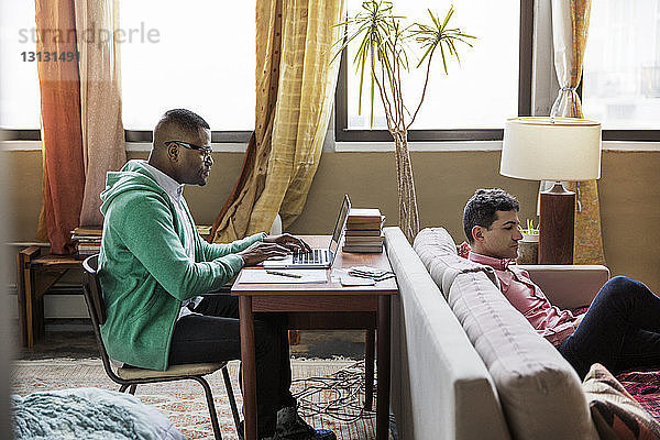 Seitenansicht eines schwulen Mannes mit Laptop am Tisch  während der Partner auf dem Sofa sitzt