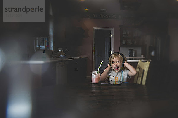 Fröhliches Mädchen hört Musik über Tablet-Computer und trinkt gleichzeitig Milchshake bei Tisch