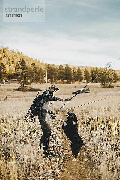 Männlicher Wanderer hält Angelruten und spielt mit Hund  der auf dem Feld gegen den Himmel steht