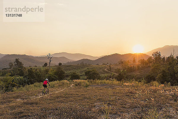 Hochwinkelansicht eines Mannes beim Mountainbiking auf dem Feld gegen den Himmel bei Sonnenuntergang