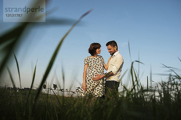 Niedrigwinkelansicht eines Mannes  der den Bauch einer PRegnantin berührt  während er auf einem Grasfeld vor klarem Himmel steht