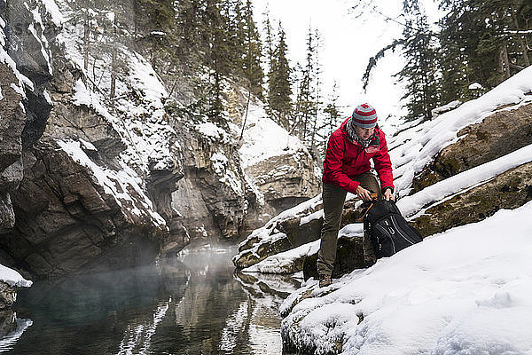 Wanderer mit Rucksack auf schneebedeckten Felsen am Bach im Wald stehend