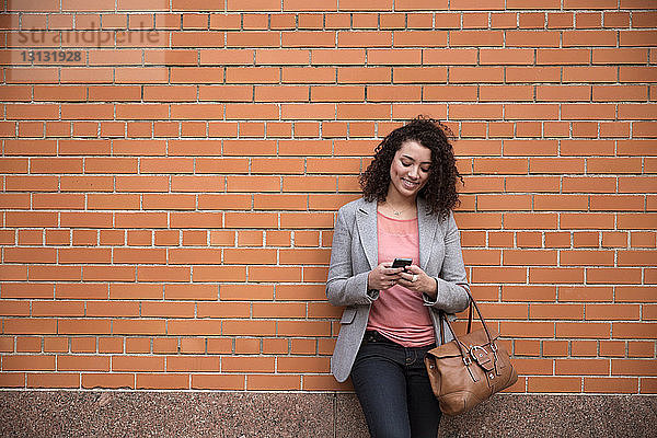 Lächelnde Geschäftsfrau benutzt Smartphone  während sie an einer Ziegelmauer steht