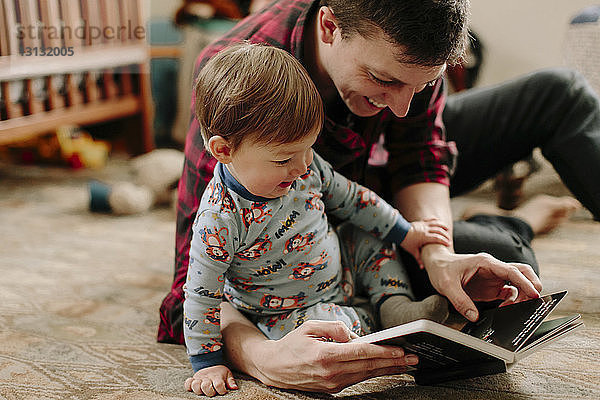 Glücklicher Vater zeigt seinem Sohn ein Bilderbuch  während er zu Hause auf einem Teppich sitzt