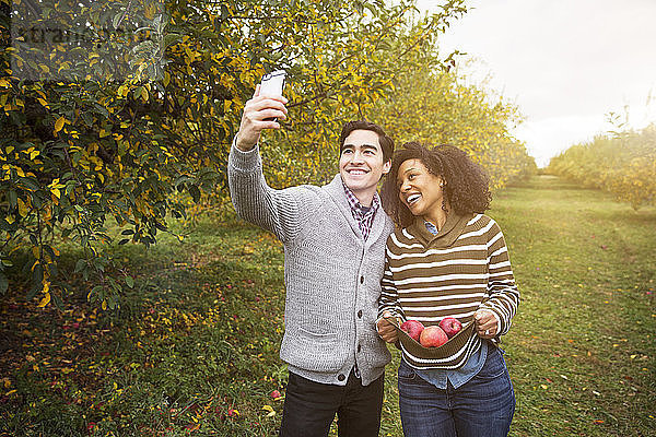 Mann nimmt sich selbst  während er mit seiner Freundin im Obstgarten steht und Äpfel trägt
