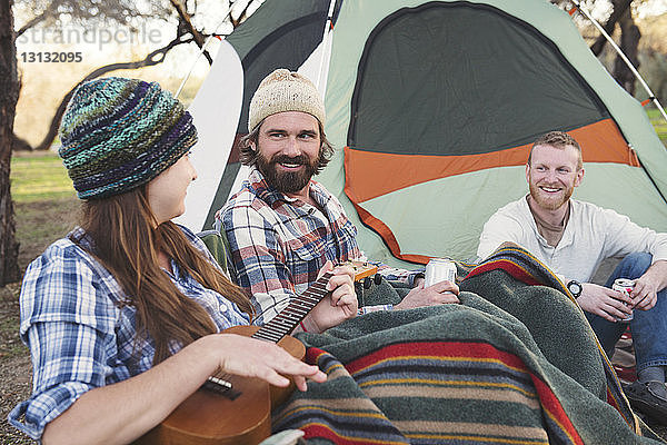 Glückliche Männer sehen eine Frau an  die vor einem Zelt im Wald Gitarre spielt