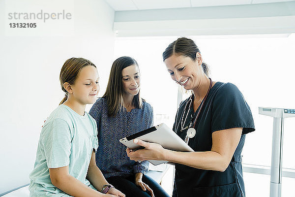 Fröhliche Kinderärztin zeigt Mutter und Tochter  die im Krankenhaus auf dem Untersuchungstisch sitzen  einen Tablet-Computer