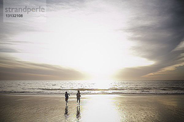 Verspielte Brüder rennen bei Sonnenuntergang am Strand gegen bewölkten Himmel