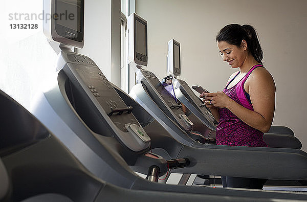 Lächelnde Frau benutzt Smartphone  während sie im Fitnessstudio auf dem Laufband steht