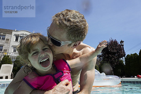 Niedrigwinkelansicht eines hemdlosen Vaters  der seine glückliche Tochter küsst  während er an einem sonnigen Tag im Schwimmbad eines Touristenortes schwimmt