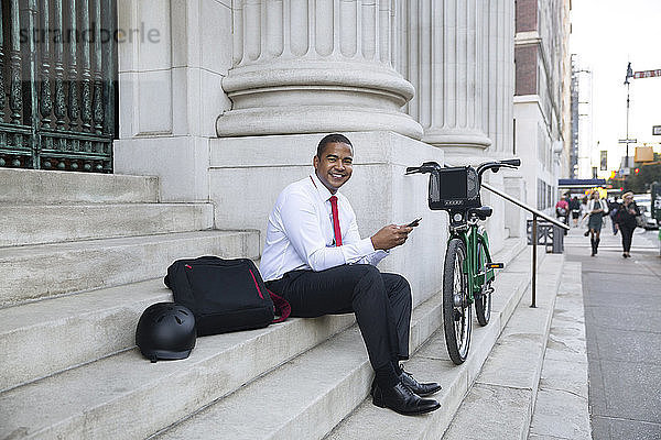 Porträt eines glücklichen Geschäftsmannes  der ein Mobiltelefon benutzt  während er auf einer Treppe sitzt