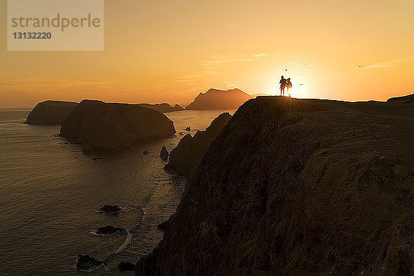 Silhouettenpaar steht bei Sonnenuntergang auf einer Felsformation am Meer