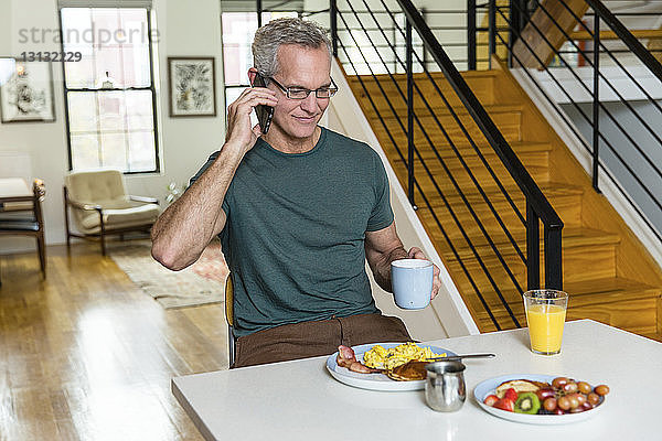 Reifer Mann hält Kaffeetasse in der Hand  während er am Esstisch an ein Smartphone geht