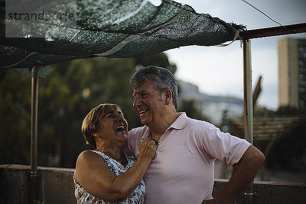 Glückliches älteres Ehepaar lacht  während es bei Sonnenuntergang auf der Gebäudeterrasse steht