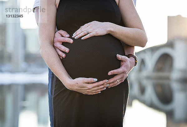 Mittelteil des Ehemannes berührt den Bauch der schwangeren Frau  während er am Fluss steht