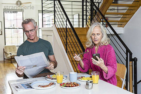 Mann liest Zeitung  während die Frau ein Smartphone am Esstisch benutzt