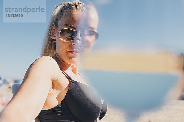 Porträt einer Frau mit Sonnenbrille und Bikini am Strand mit einem Trinkglas im Vordergrund