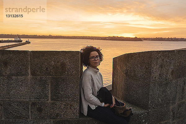 Porträt einer Frau  die bei Sonnenuntergang auf der Stützmauer eines historischen Gebäudes gegen das Meer sitzt