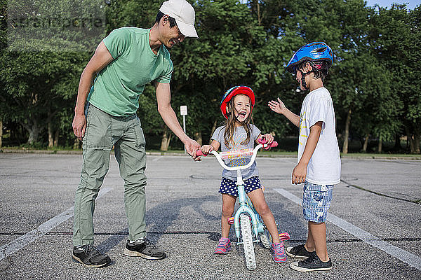 Fröhliches Mädchen lernt Radfahren mit Vater und Bruder auf dem Spielplatz