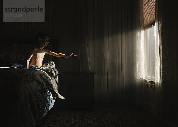 Junge ohne Hemd  der versucht  das Sonnenlicht zu berühren  während er in der Dunkelkammer auf dem Bett sitzt